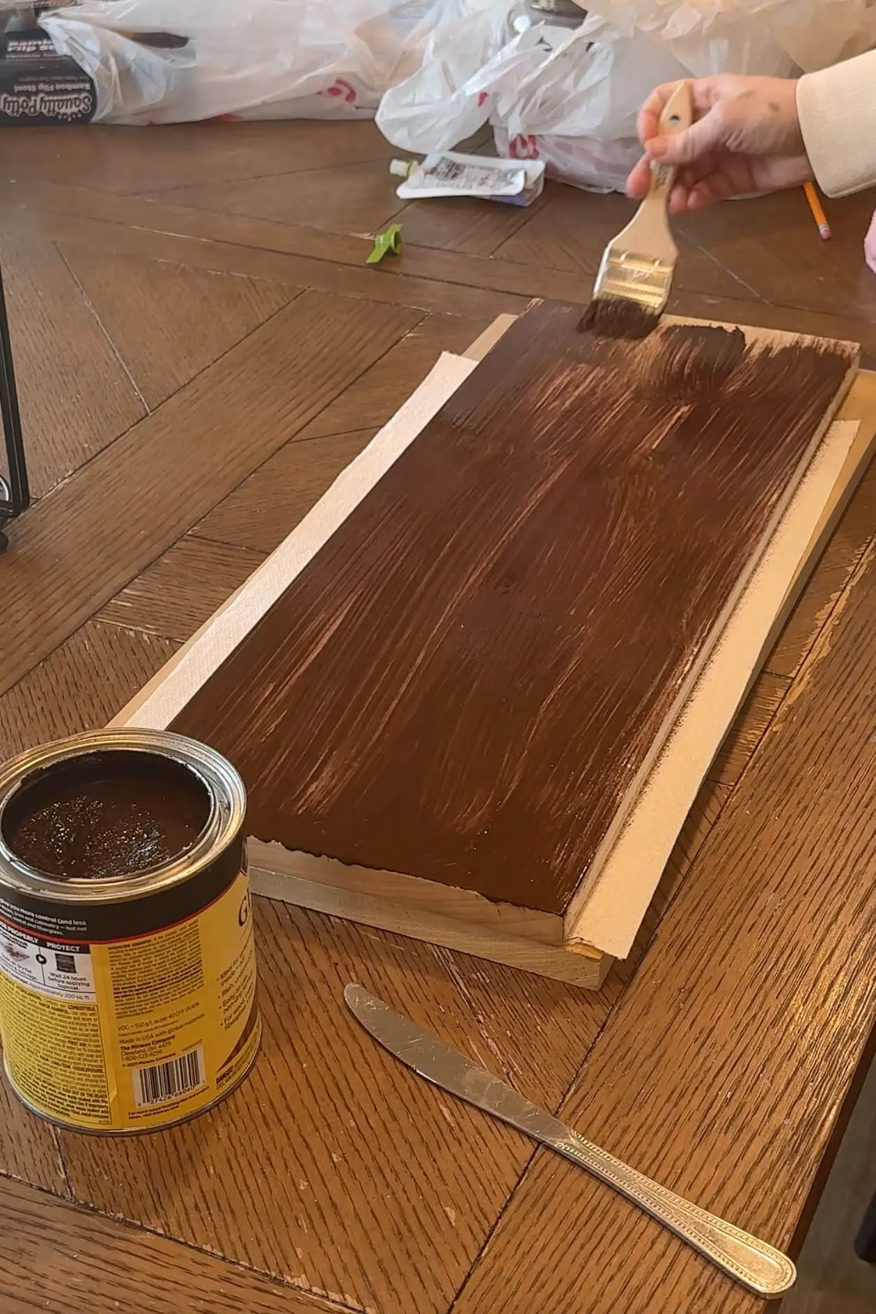 A piece of raw wood being stained with Minwax Early American gel stain, with a brush and open can of stain in the foreground.